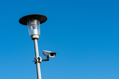 A streetlight stands tall, featuring a surveillance camera attached to its pole. The backdrop is a...