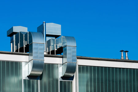 Metal ventilation pipes and ducts are seen on a modern building against a clear blue sky. The structure features glass panels, showcasing contemporary architectural design.の写真素材