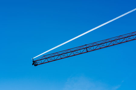 A construction crane rises against a vivid blue sky, where a white contrast from a passing aircraft streaks across the scene.の写真素材