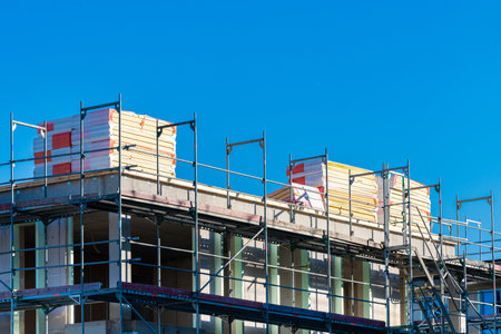 A building under construction surrounded by scaffolding. Building materials in packaging under blue...
