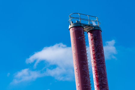 Two tall, weathered chimneys stood prominently against a bright blue sky. The structures show signs of age, with peeling paint highlighting their industrial character in a sunny environment.の写真素材