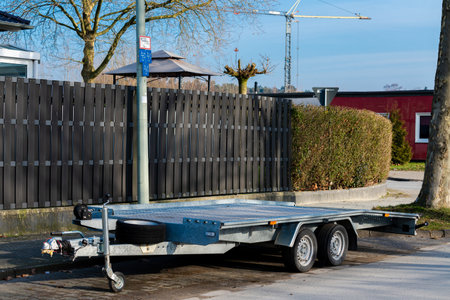 A metallic trailer is parked next to a hedge, with construction cranes visible in the background....