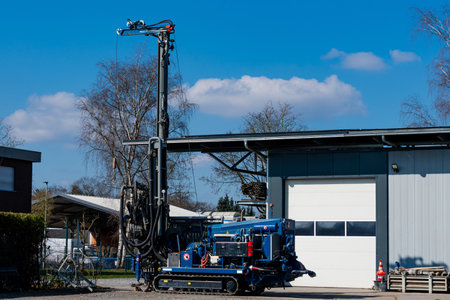 Heavy drilling equipment stands next to an industrial building under a bright blue sky. The...