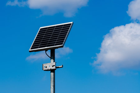 A solar panel stands tall on a metal pole amidst a clear blue sky with fluffy white clouds. This...