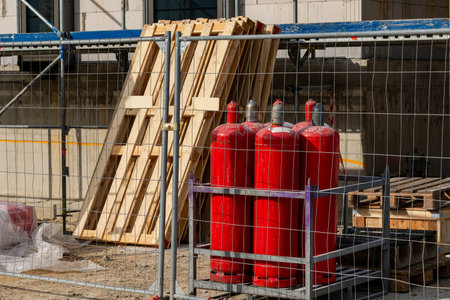 Red gas cylinders are secured behind a metal fence at a construction site, while wooden pallets lean...