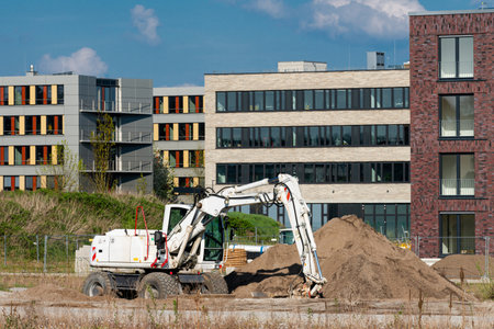 An excavator digs into a large mound of sand at a construction site near residential buildings....