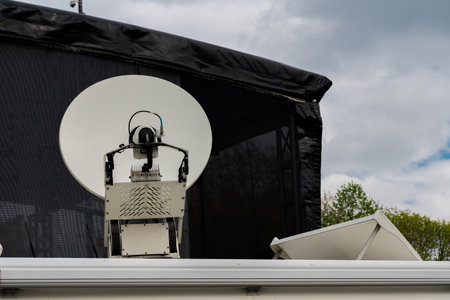 A large satellite dish is positioned on a rooftop, with equipment visible for transmission. The background features a dark cover and a cloudy sky, indicating potential rain.の写真素材