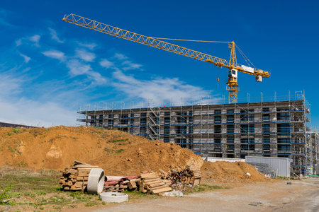 A large construction site features a towering crane above a partially built structure surrounded by scaffolding. Nearby, stacks of wood and piles of dirt lie under a vibrant blue sky.の写真素材