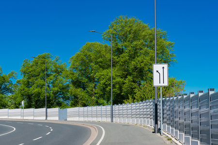 A curved roadway features directional signs pointing ahead and to the left. Lush green trees line the road, creating a vibrant contrast against the clear blue sky on a bright day.の写真素材