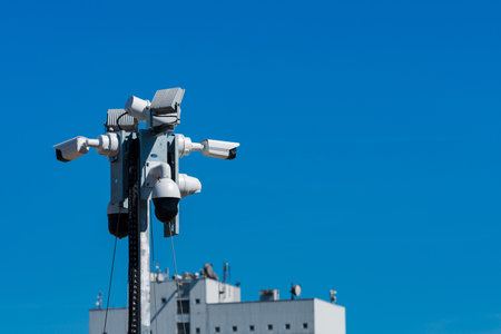 A collection of surveillance cameras mounted on a tall pole overlooks a building. The cloudless blue sky creates a stark background, emphasizing the focus on security equipment.の写真素材
