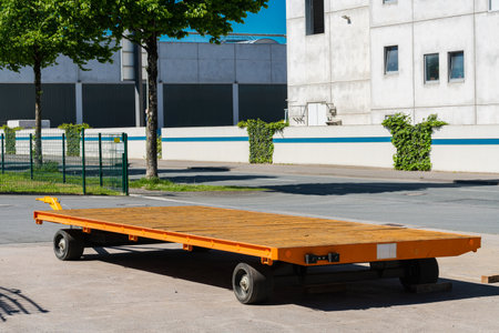 A large, orange industrial transport trailer is parked on the pavement outside a building. Trees provide shade, and bright sunlight creates a vivid atmosphere in the urban environment.の写真素材