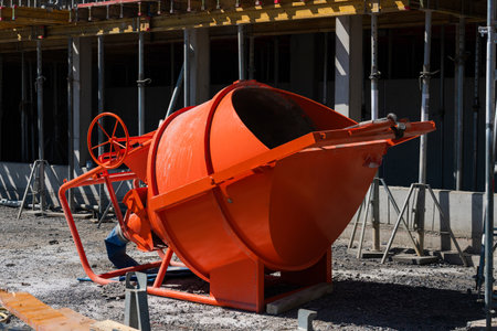 Workers are using an orange concrete mixer at a construction site. The mixer is positioned on a gravel surface, surrounded by scaffolding and building materials. It's a sunny day.の写真素材