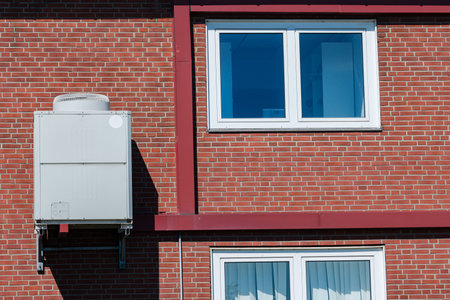 An air conditioning unit is fixed to a brick wall above two windows. The bright sunlight casts shadows, highlighting the building's structure and design.の写真素材