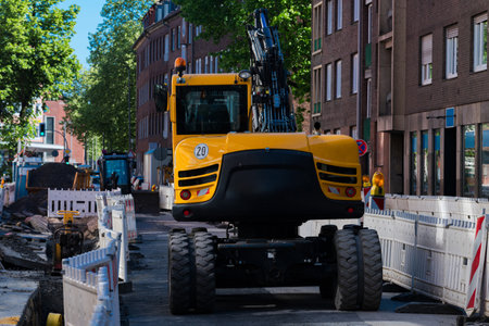 A yellow excavator is positioned on a city street among construction barriers. Nearby workers manage...