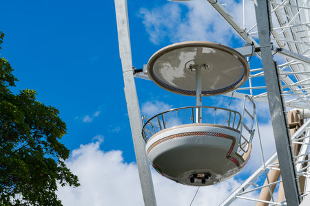 A single gondola of a Ferris wheel hangs against a vibrant blue sky dotted with fluffy white clouds. The structure is partly shaded by a nearby tree, enhancing the scene's brightness.の写真素材