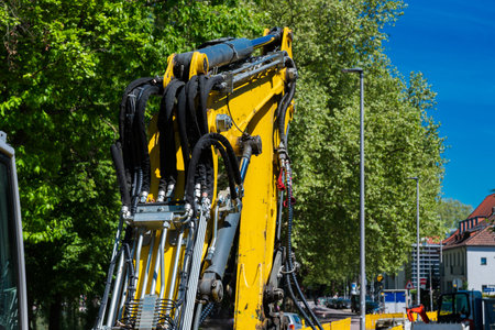 A yellow excavator is parked on a city street, surrounded by lush green trees. The machine's arm is positioned upwards, indicating preparation for a task.の写真素材