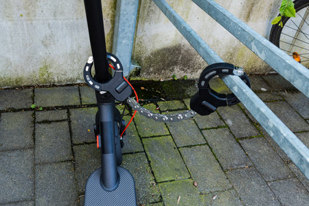 An electric scooter is securely fastened to a bike rack using a metal lock chain. The setting is an urban environment with a concrete backdrop and some greenery visible nearby.の写真素材