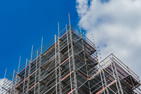 Tall scaffolding is displayed against a clear blue sky dotted with white clouds. The structure is...