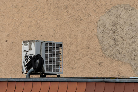 An air conditioning unit sits atop a building's roof, surrounded by a pale wall. The device is partially shaded, indicating a warm day in an urban environment.の写真素材