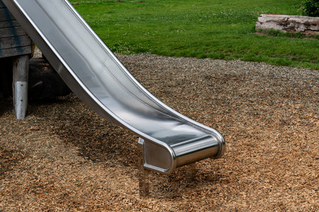 A shiny stainless steel slide stands at a playground surrounded by grass and wood chips. The outdoor space is sunny and inviting, perfect for recreation and leisure activities.の写真素材
