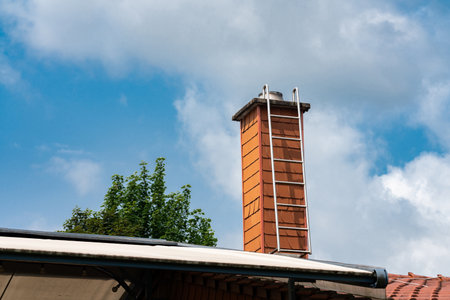 A tall red brick chimney rises against a backdrop of bright blue sky and fluffy clouds. Lush green trees surround the structure, indicating a residential environment.の写真素材