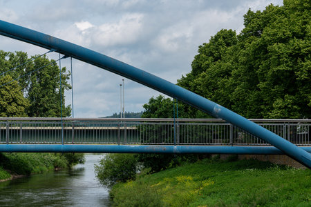 A modern bridge stretches across a calm river, surrounded by lush greenery and trees. Overcast skies create a serene backdrop for the peaceful urban landscape by the water's edge.の写真素材