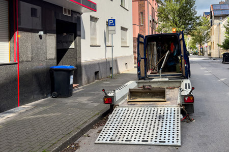 A delivery truck is parked with its ramp lowered on a city street, showing equipment inside. Nearby, there is a trash bin and buildings lining the street on a sunny afternoon.の写真素材