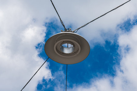 A round light fixture hangs from overhead wires, set against a vibrant blue sky filled with fluffy white clouds. The fixture adds a sense of modernity to the outdoor space below.の写真素材