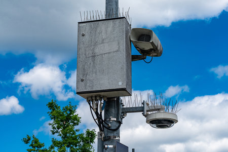 A concrete surveillance box sits on a tall pole alongside a camera and speakers. Spiky deterrents are placed around the equipment to prevent tampering. Fluffy clouds are visible above.の写真素材