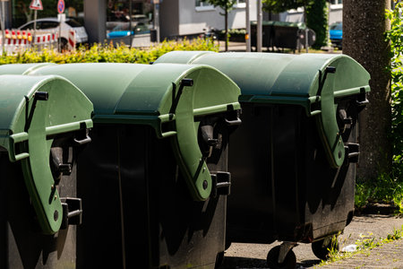 A group of green trash bins is lined up on the sidewalk of an urban area. The sun shines brightly, illuminating the bins and surrounding greenery. City buildings are visible in the background.の写真素材