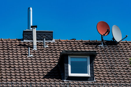 Bright sunlight highlights a rooftop with satellite antennas and a chimney. The clear blue sky serves as a backdrop, accentuating the unique features and textures of the roof.の写真素材