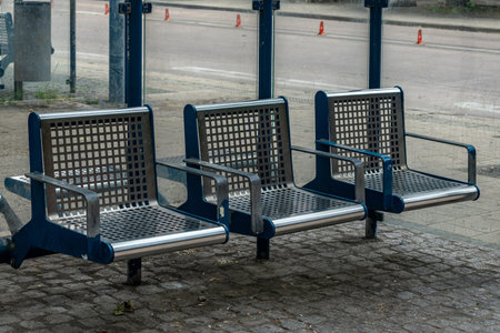 Three modern metal benches are positioned at a bus stop in an urban area. The benches feature a grid design and are set on a paved surface, ready for waiting passengers.の写真素材