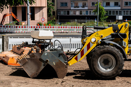 A yellow excavator is seen operating on a construction site in a city environment. The machine is digging into the ground while other equipment is visible in the background under clear blue skies.の写真素材