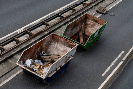 Two large dumpsters sit alongside a highway, filled with construction debris and waste materials. The setting features a gray road and nearby construction elements, indicating active work.の写真素材