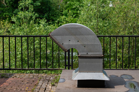 A unique metal ventilation structure is positioned next to a calm body of water, framed by lush...