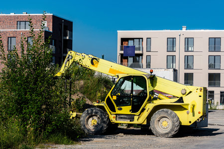 A yellow telehandler is working on a construction site surrounded by modern buildings. Vegetation is...