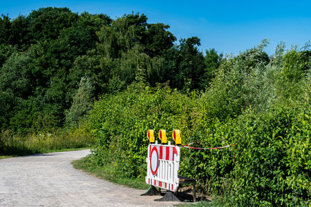 A construction barrier with stop signal stands on a gravel path in a vibrant green landscape. Tall trees and dense foliage surround the area under a clear blue sky.の写真素材