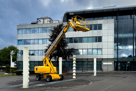 A bright yellow lift is raised near a contemporary office structure, showcasing its height and work...