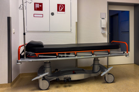 A wheeled stretcher stands ready in a hospital corridor. The stretcher features a padded surface and is equipped for patient transport. Nearby, safety signs indicate important hospital protocols.の写真素材