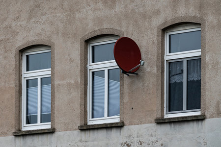 A red satellite dish is attached to the exterior wall of a building. Three white-framed windows can be seen nearby, creating a simple urban scene. The dish is positioned for optimal signal reception.の写真素材