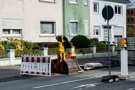 Heavy machinery is parked on the road in front of residential houses. A construction site with barriers can be seen, indicating ongoing street work during daylight hours.の写真素材