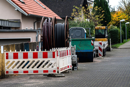 Heavy machinery and construction barriers are seen on a street in a residential neighborhood. Cable reels are placed nearby, indicating ongoing utility work.の写真素材