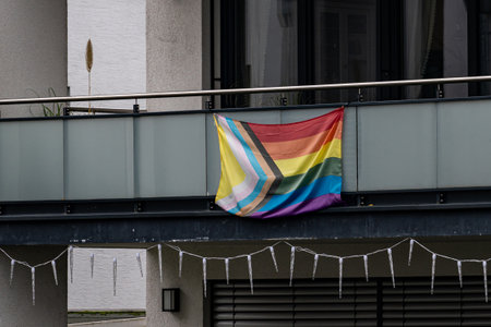 A colorful rainbow flag hangs from a balcony in an urban area, symbolizing LGBTQ+ pride and...