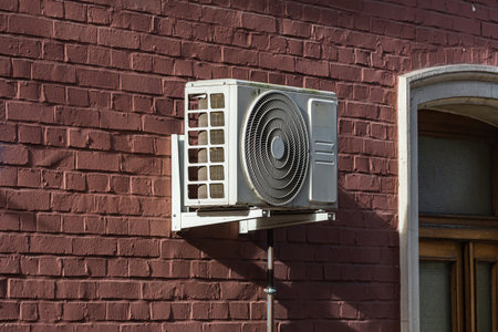 An air conditioning unit is mounted on a red brick wall outside a building. The unit is positioned beneath a window, providing cooling for the indoor space during warm weather.の写真素材