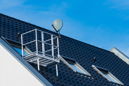 A metal maintenance platform is positioned on a roof alongside a satellite dish. The clear blue sky provides a bright backdrop, highlighting the residential building and its unique roofing details.の写真素材