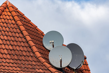 Three satellite dishes are mounted on a red tiled roof. The sky is partly cloudy, creating a beautiful contrast with the bright tiles.の写真素材