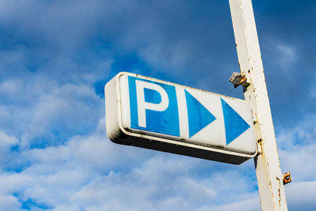 A parking sign featuring a large 'P' and two directional arrows is displayed against a backdrop of blue clouds. The sign stands tall with clear visibility, indicating parking availability.の写真素材