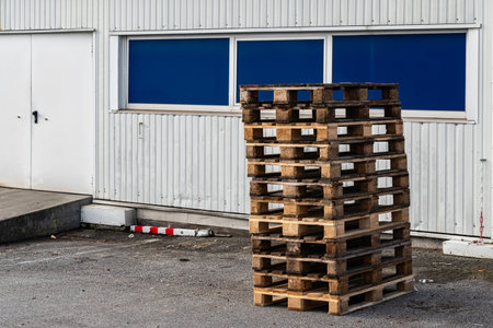 A stack of wooden pallets is positioned outside a building. The pallets are arranged neatly on gray...