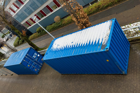 Two blue containers sit next to each other on a street. Snow covers the top of one container. Trees...