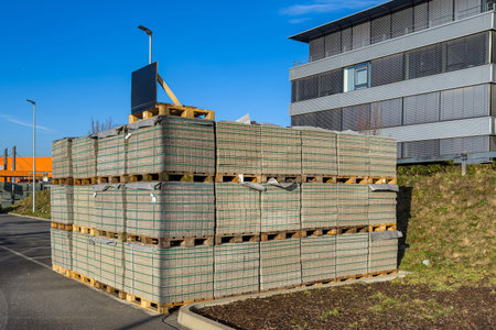 Large stacks of pallets are filled with new paving slabs. They are arranged on a paved area near a modern building under a clear blue sky. The setting shows an active construction site.の写真素材